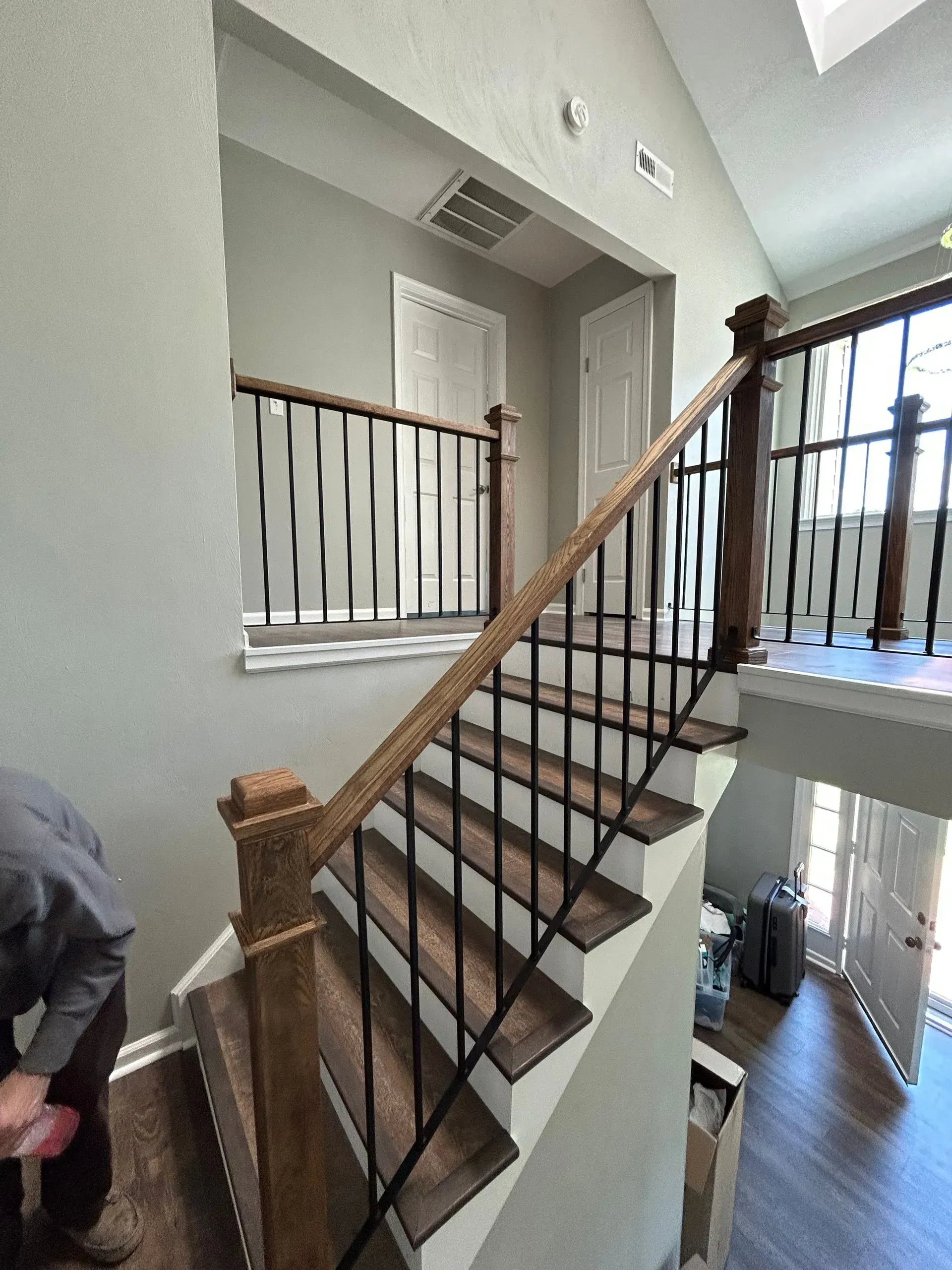 A staircase with wooden steps, railing, and posts leads up to a second-floor hallway with black metal balusters. 