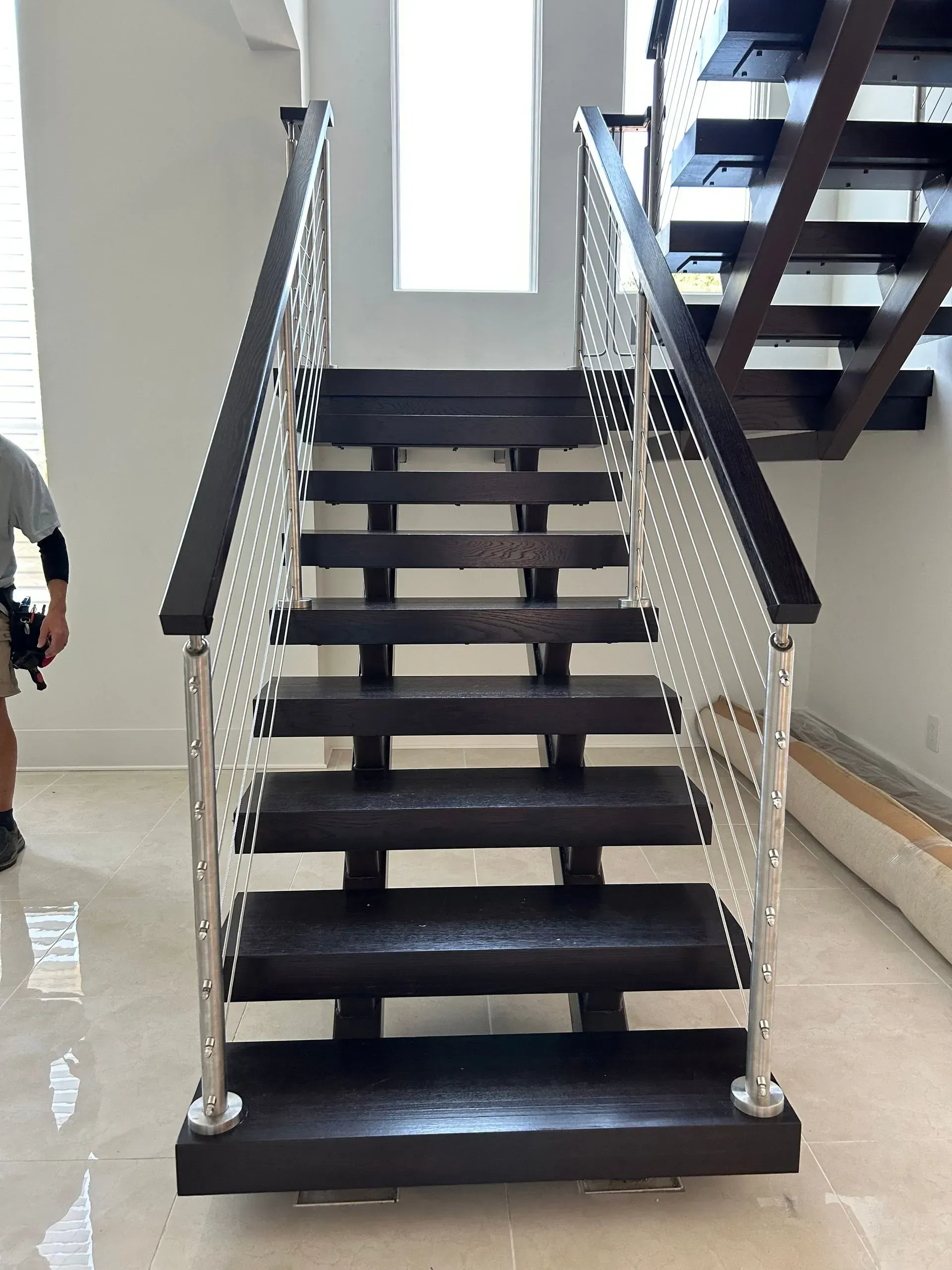 Dark wooden staircase with steel cable railings, viewed from below. A person stands to the left.