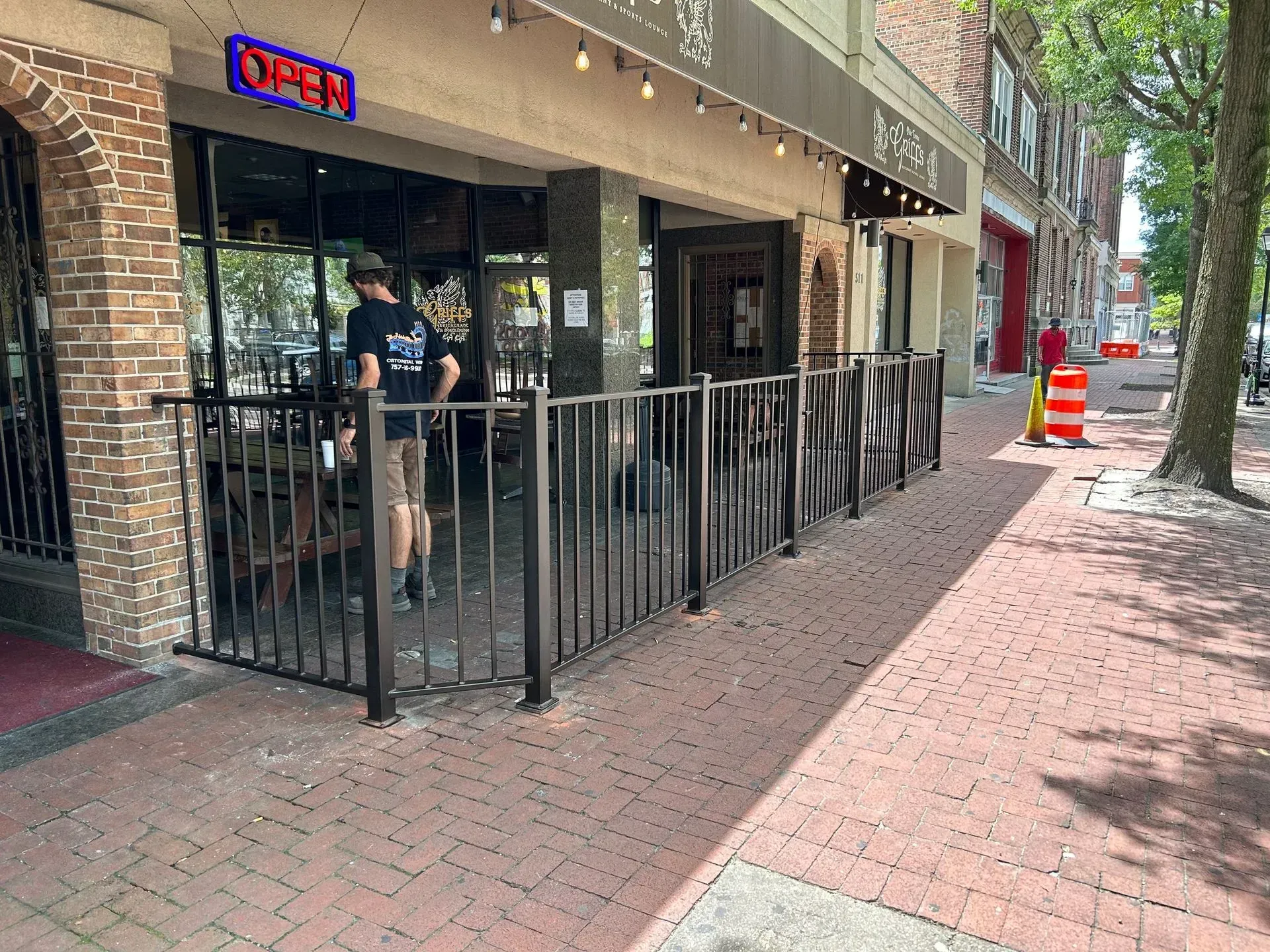 A restaurant with outdoor seating on a brick sidewalk. A person is standing inside the fenced-in area. 