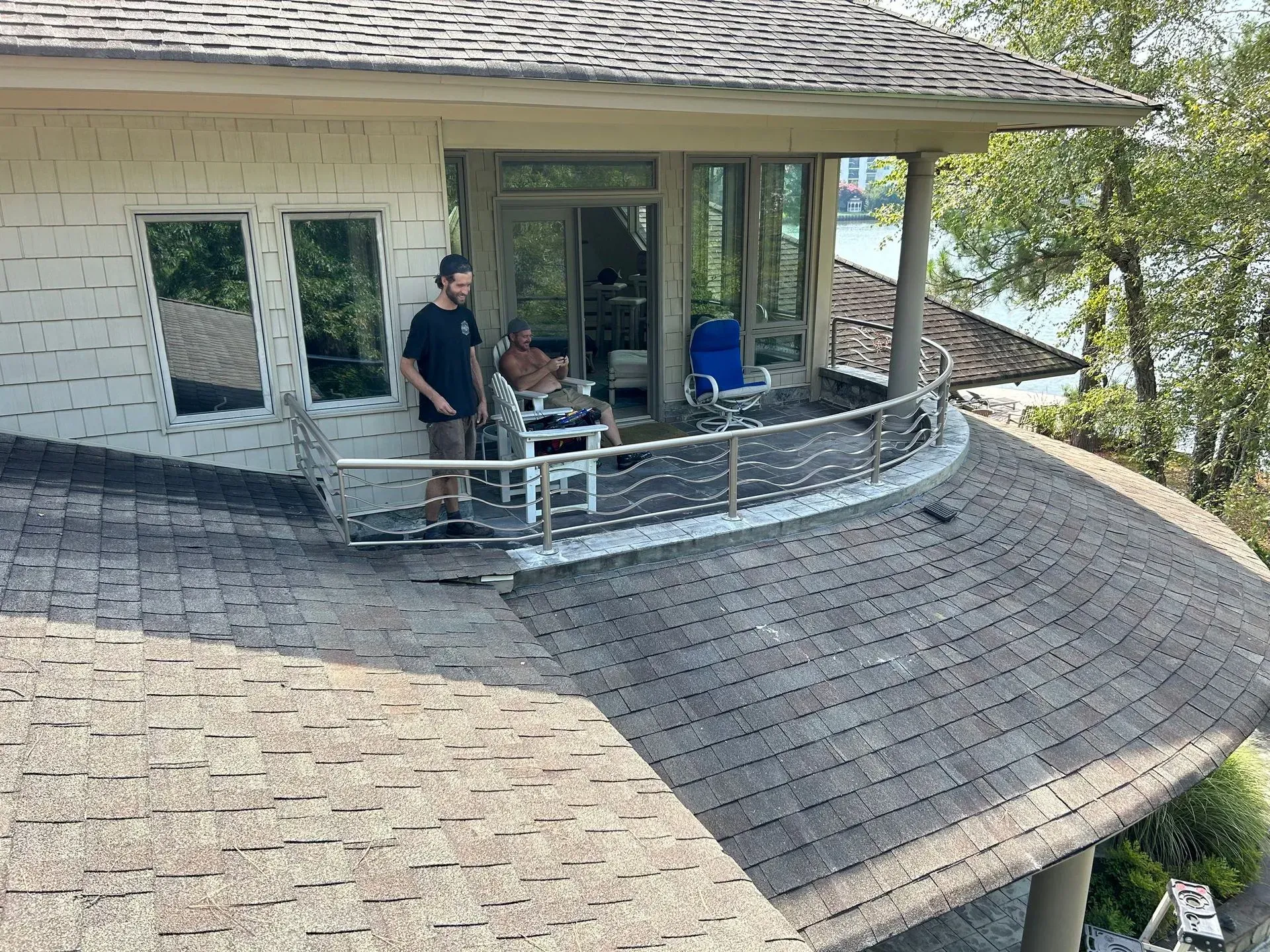 Two men on a round rooftop balcony. One stands near a railing, the other sits in a chair. Building with a lake view.