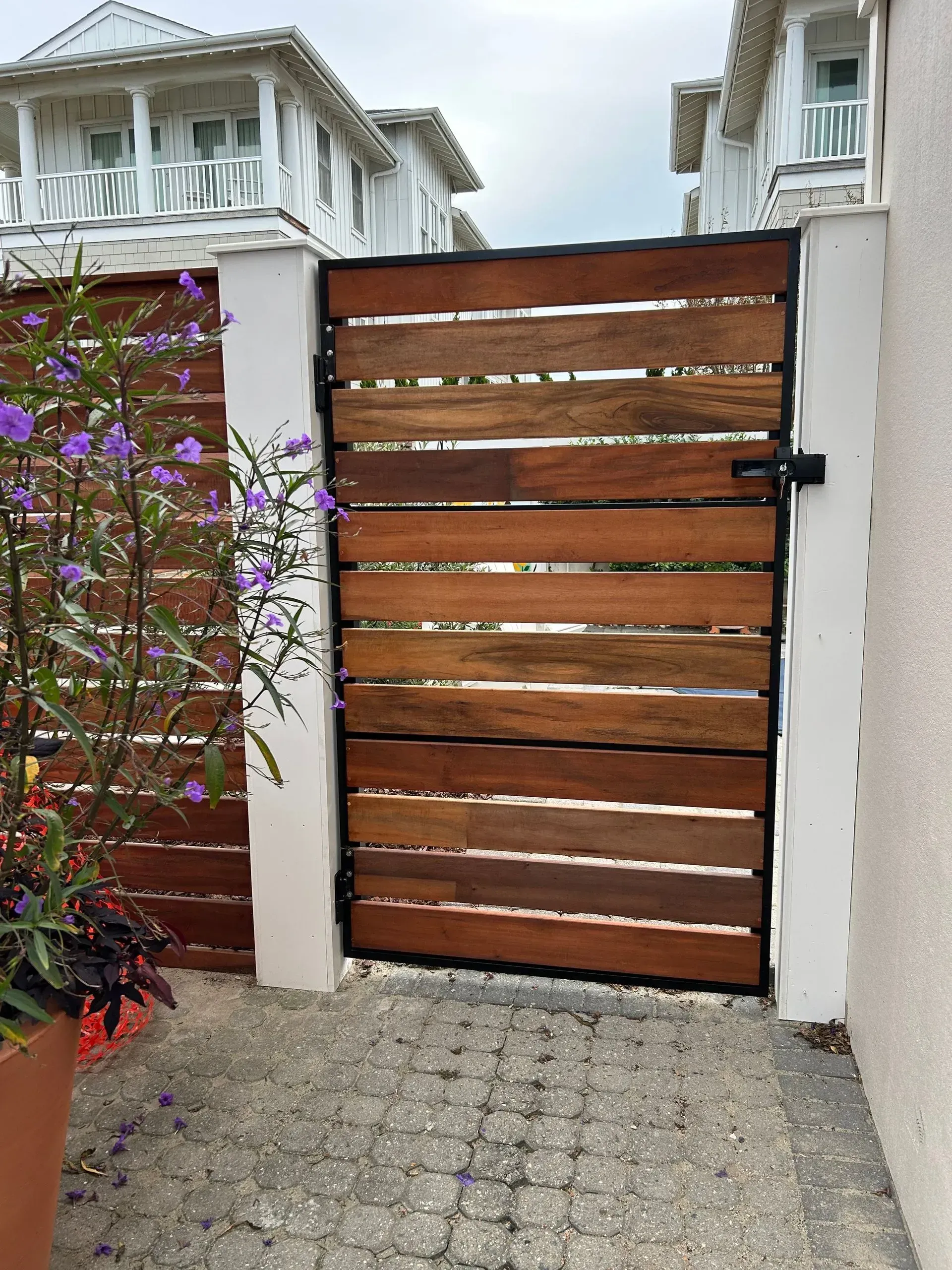 Wooden gate with dark metal frame between white pillars, leading to a pathway, with a similar fence to the left.