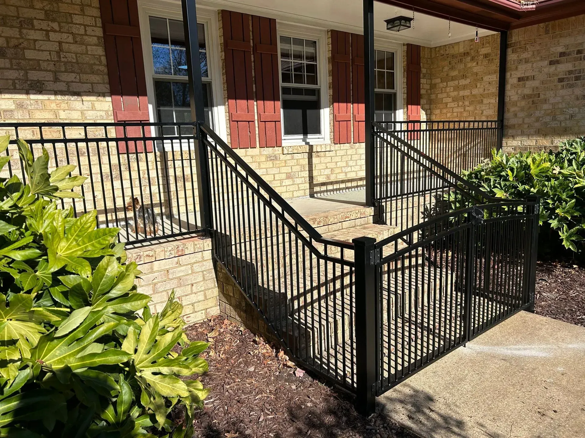 Black metal railing on the steps leading to a brick building with red shutters. Green bushes frame the stairs.