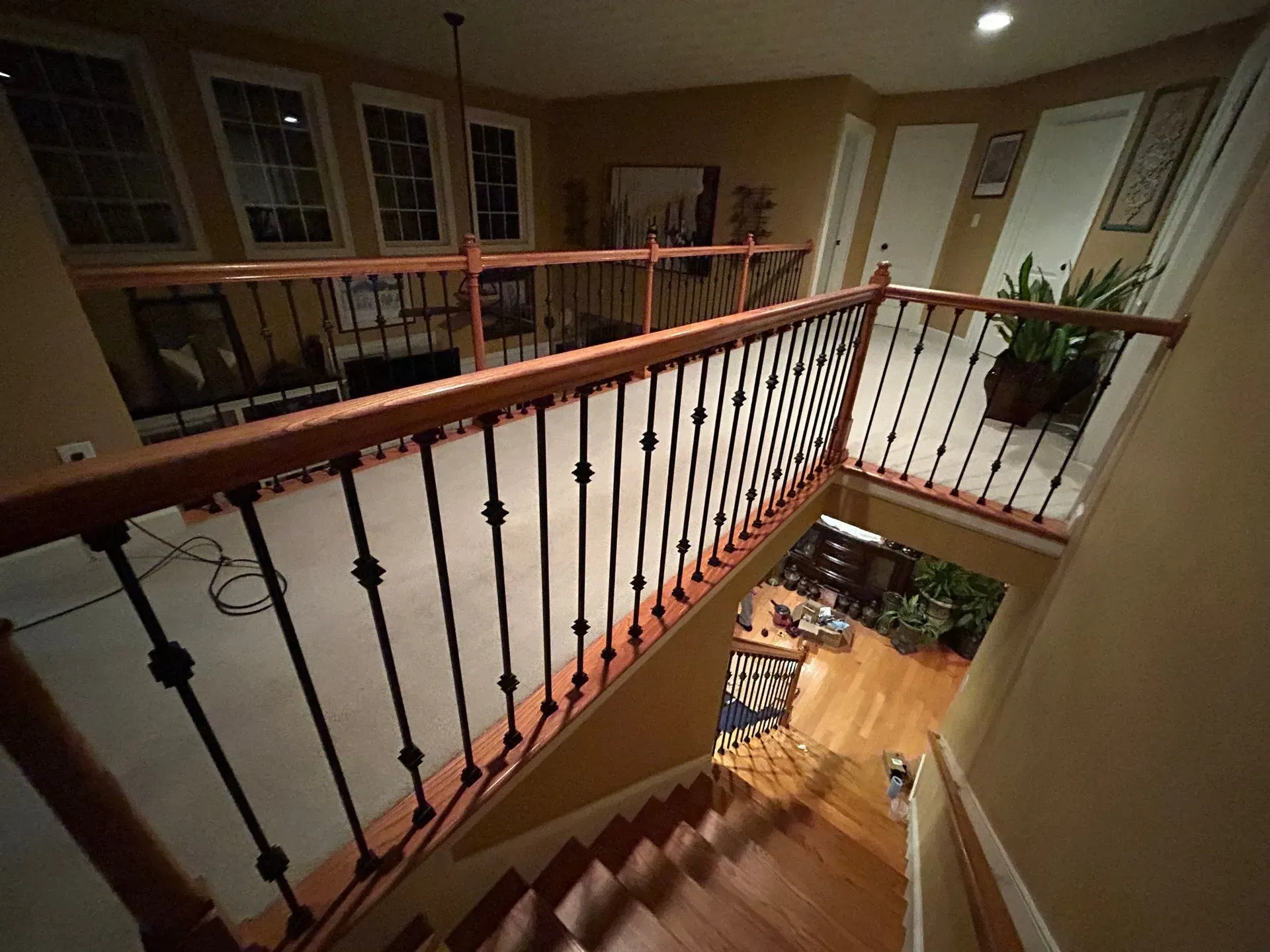 A staircase with a black railing and wooden handrail leads down into a house. A second-floor railing overlooks the stairwell.