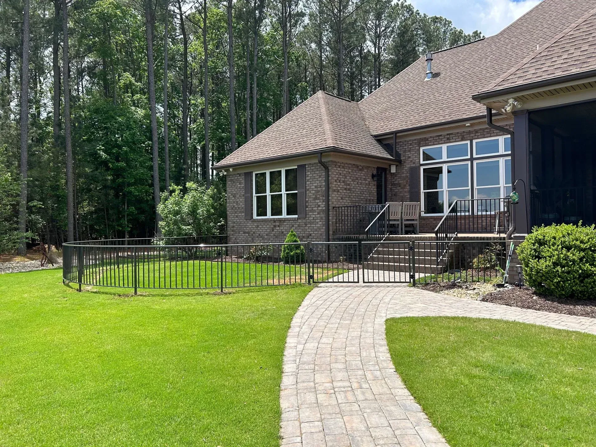 A brick house with a paved walkway leading to a black metal fenced yard and entrance. Green grass surrounds the walkway.