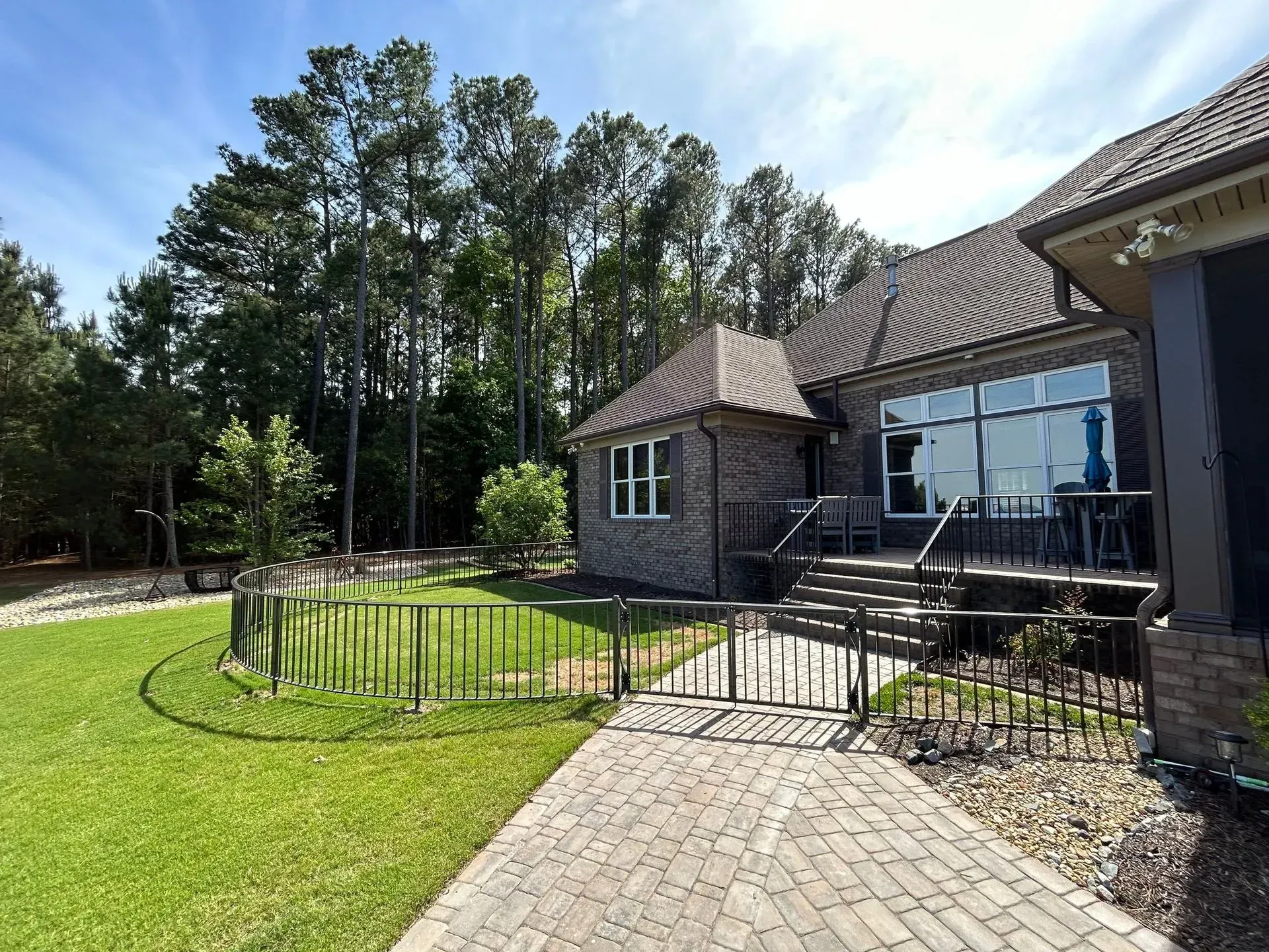 Exterior view of a brick house with a patio, green lawn, and surrounding trees on a sunny day.