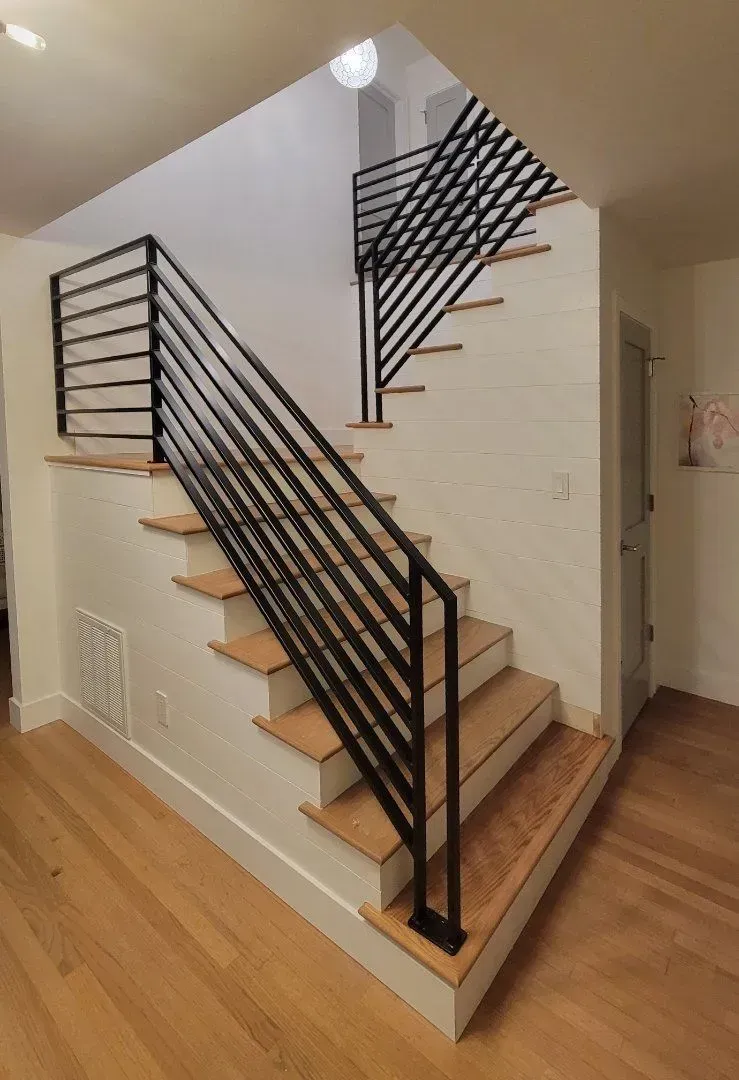 Interior view of a staircase with black horizontal railings, wooden steps, and light-colored walls and flooring.