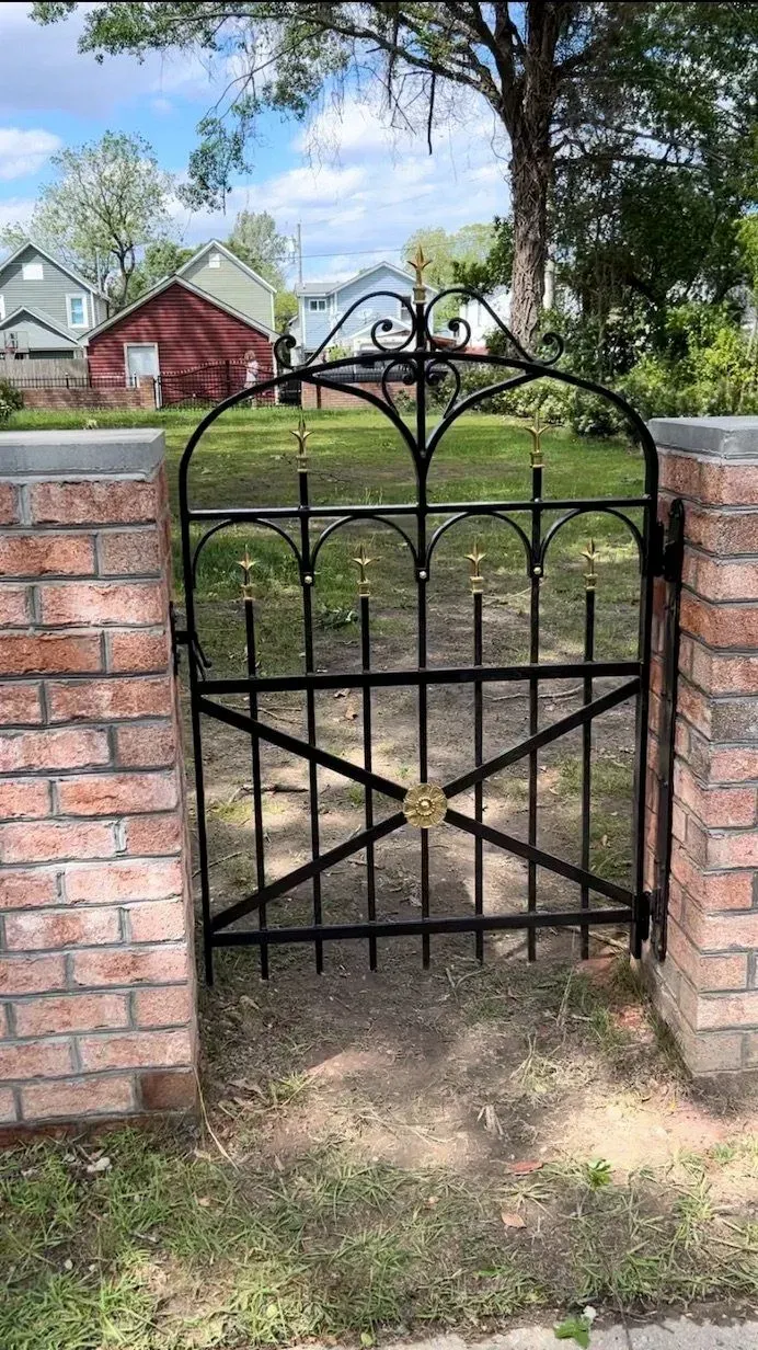 Black wrought iron gate with gold accents set between brick columns; lawn and houses visible in the background.