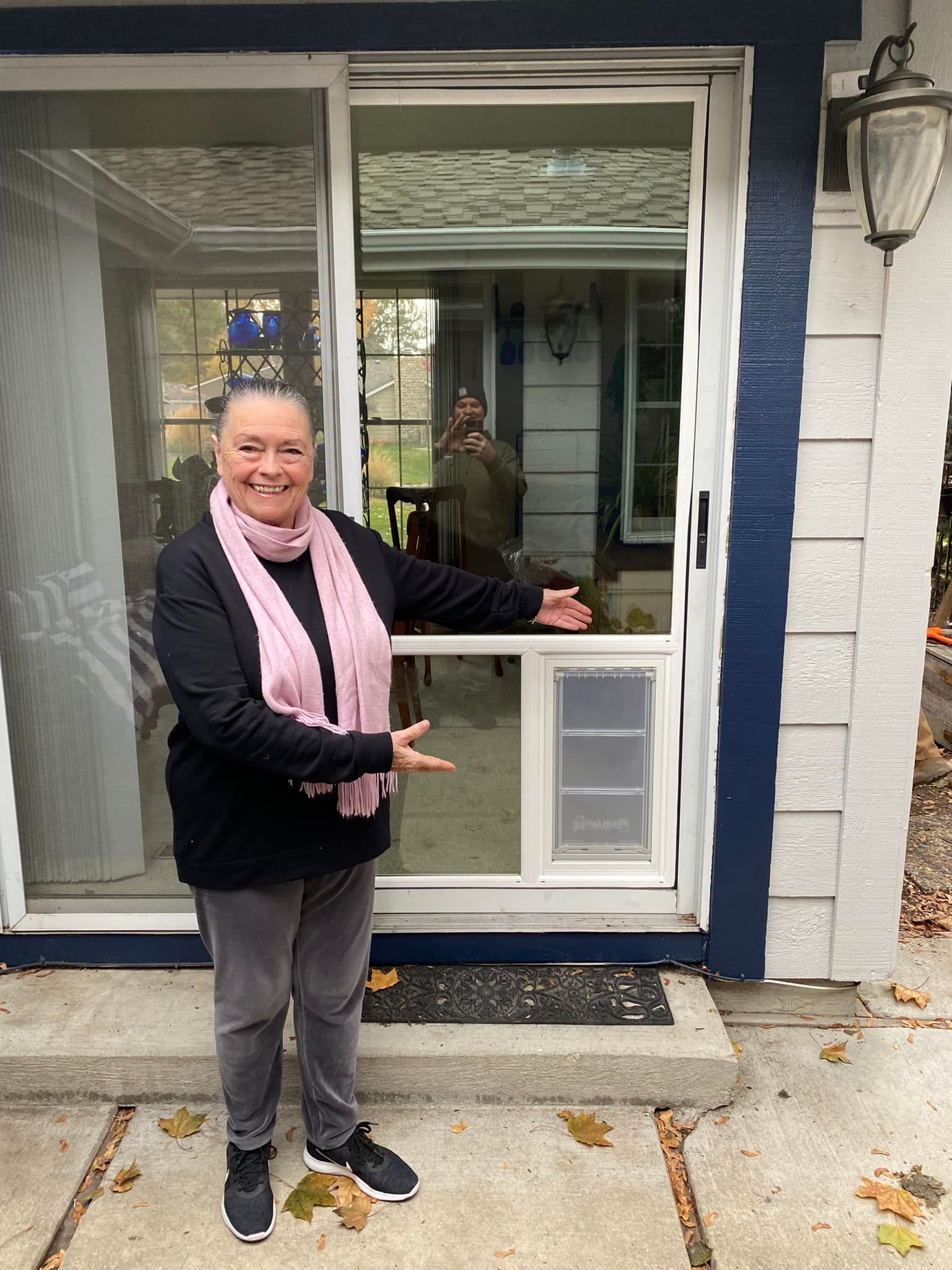 Woman points to a dog door in a window. She wears black and pink and stands outside a blue house.