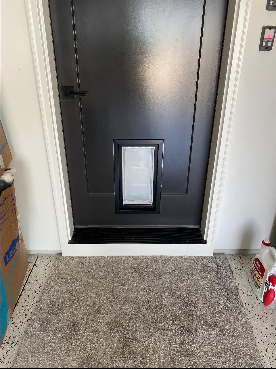 Black door with doggy door, white trim, and a gray rug on a speckled floor.