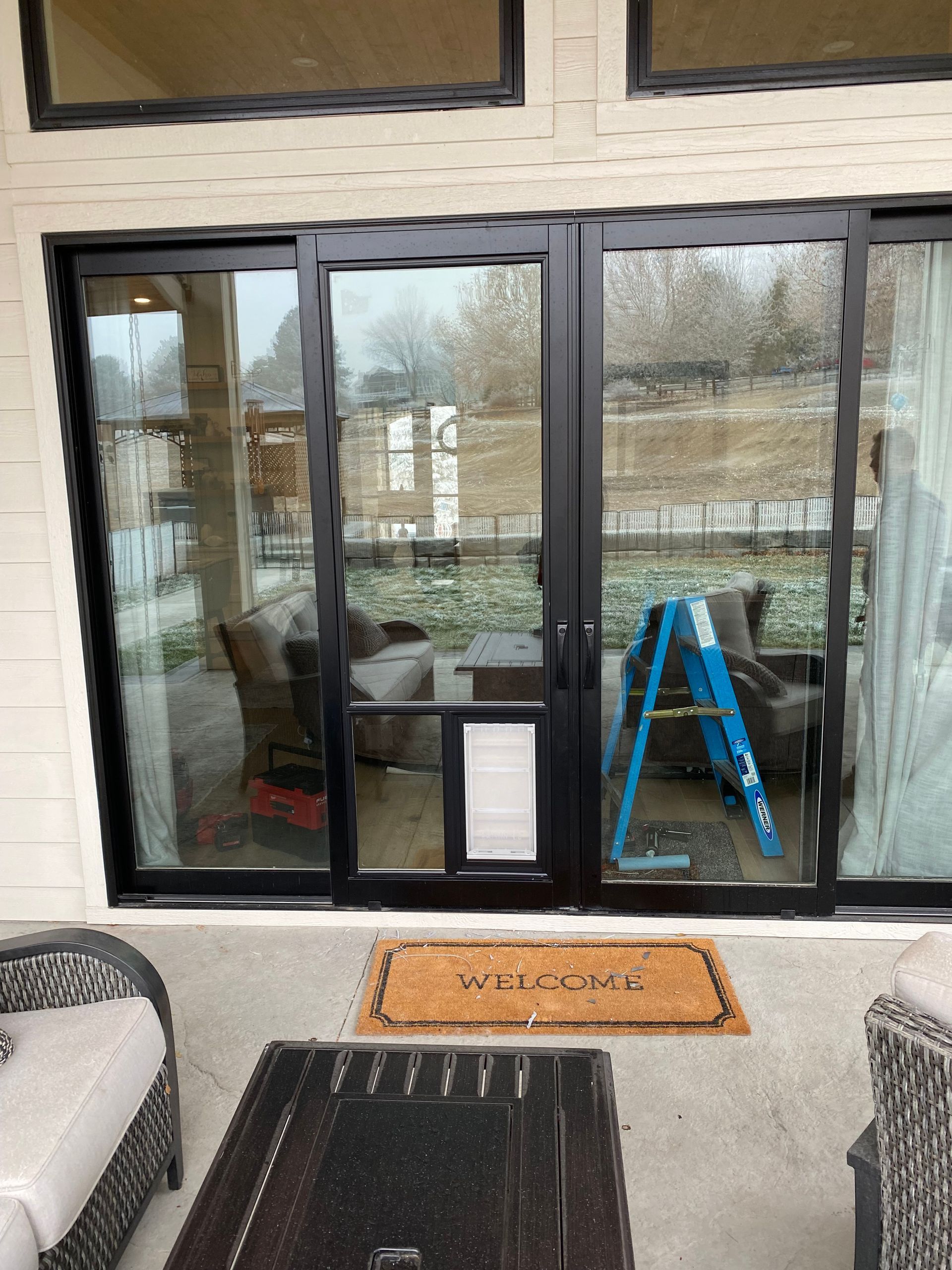 Black framed sliding glass doors with a dog door on a patio. Includes furniture and a ladder.