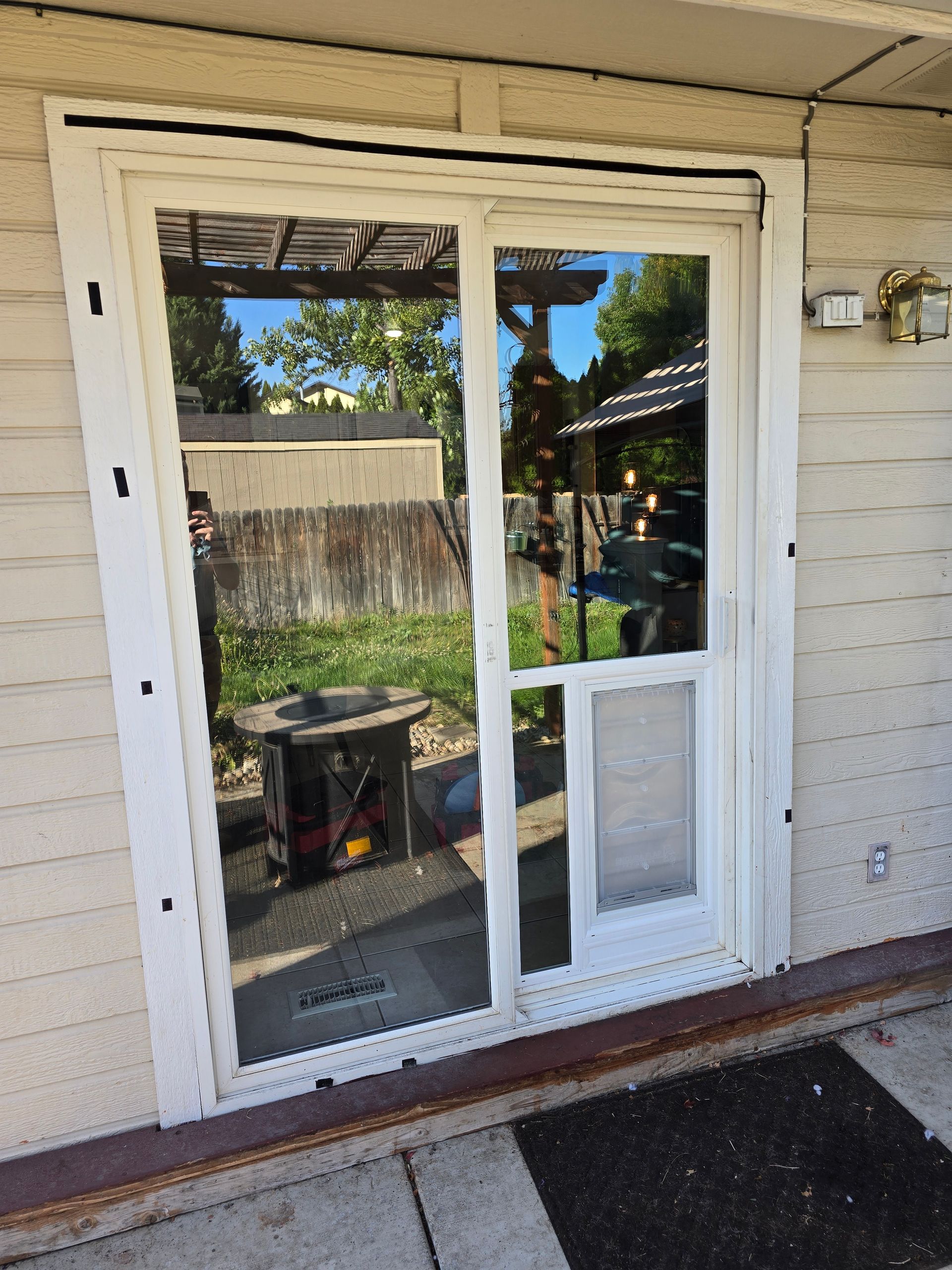 Sliding glass door with built-in dog door, reflecting a backyard with a pergola and fire pit.