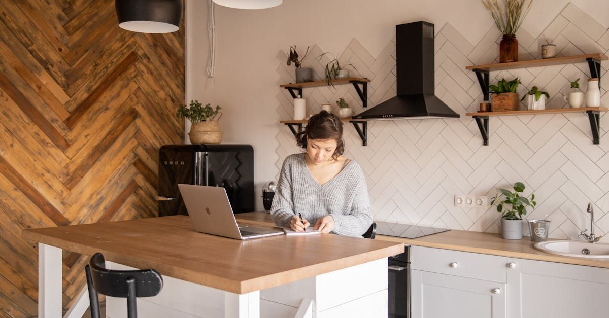 A woman is sitting at a table in a kitchen using a laptop computer.