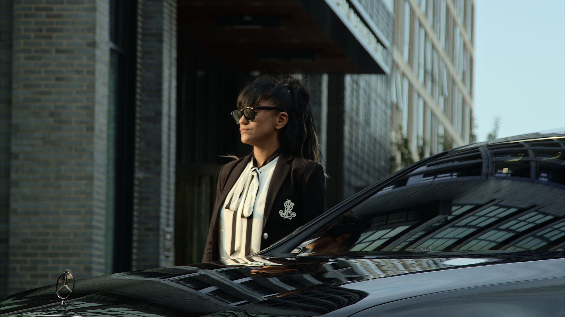 Person in sunglasses stands beside a black car on a city street.