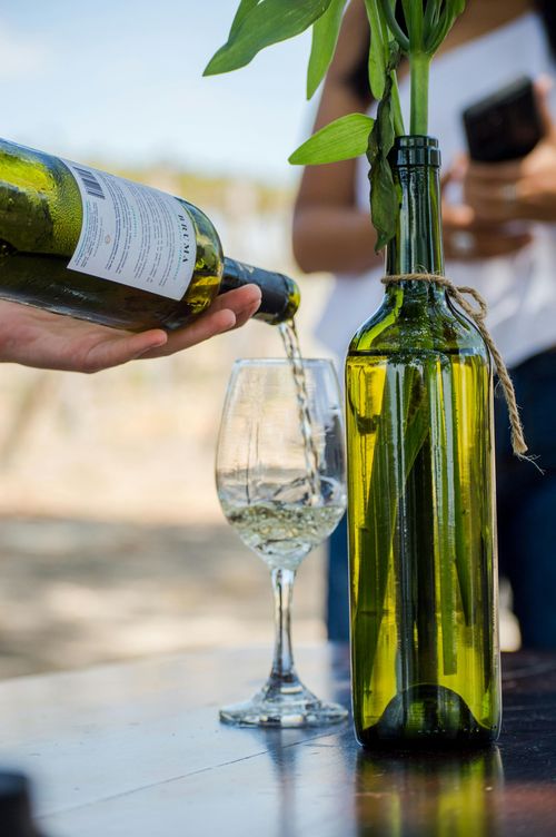 A bottle of white wine being poured into a glass beside a green bottle on a table outdoors.