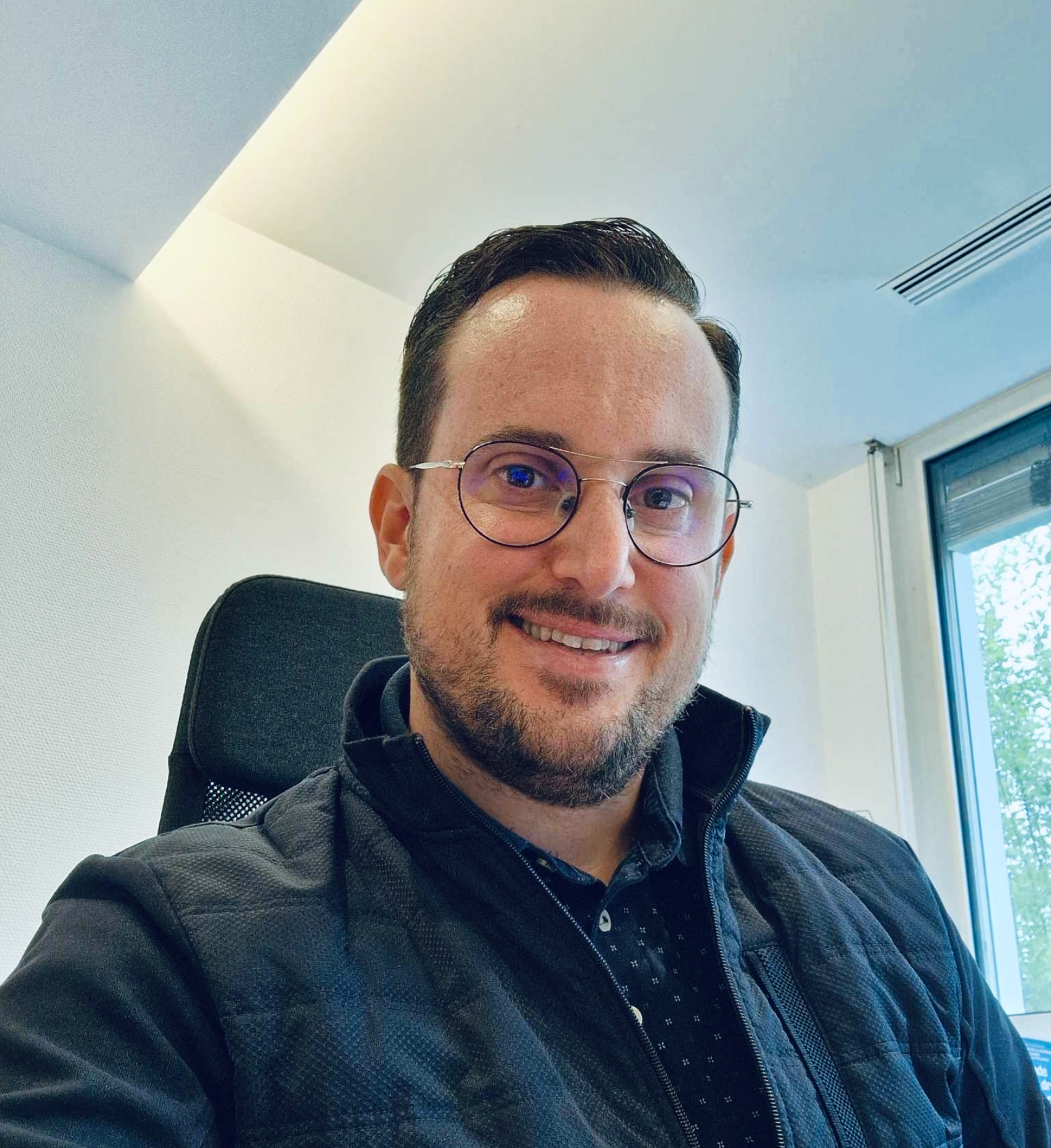 Un homme portant des lunettes et une veste noire sourit à la caméra.