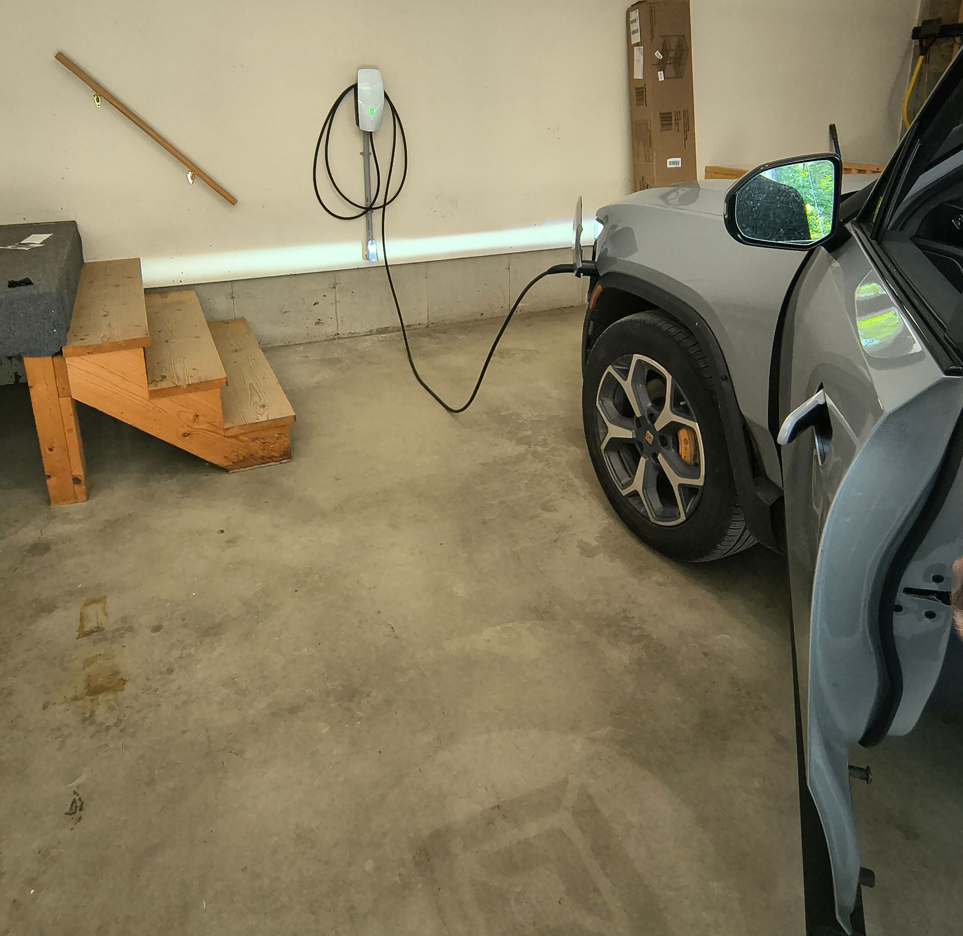 A silver electric vehicle charging in a garage. Door open, cable connected, with stairs in the foreground.