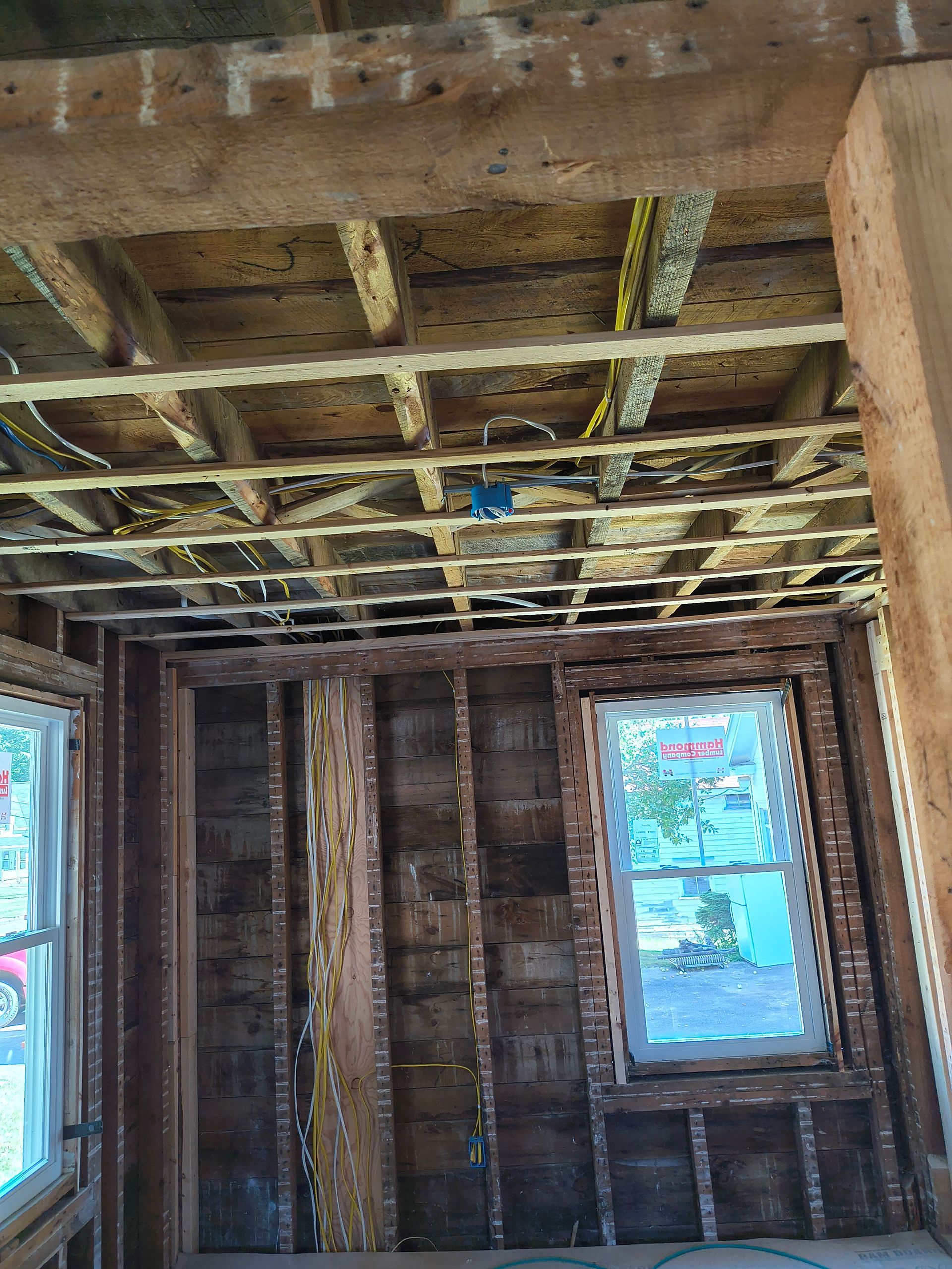 Interior of a room under construction, showing exposed wooden beams, studs, and window frames.