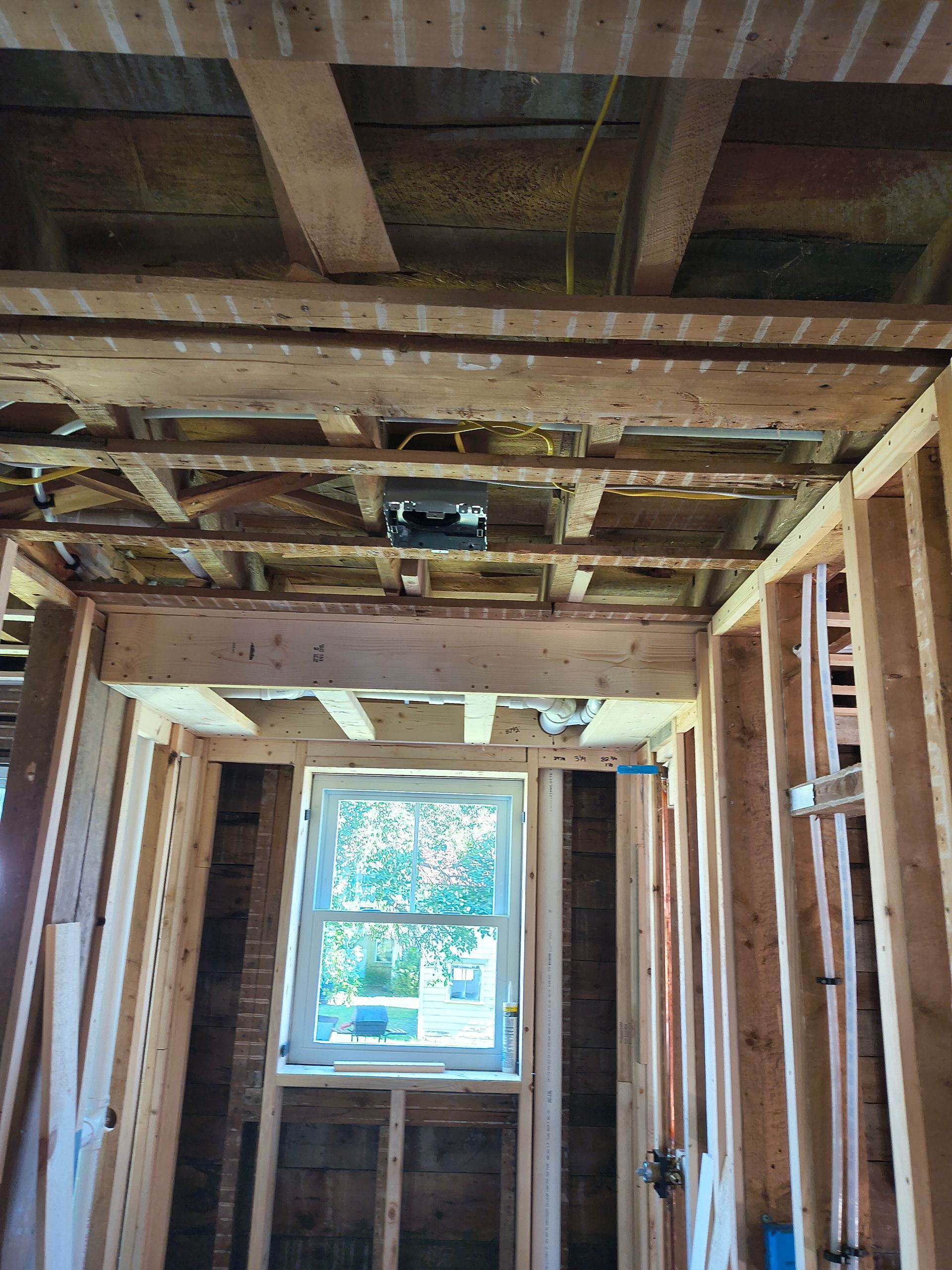 Interior view of a room under construction, with exposed beams, wood framing, window, and electrical wiring.