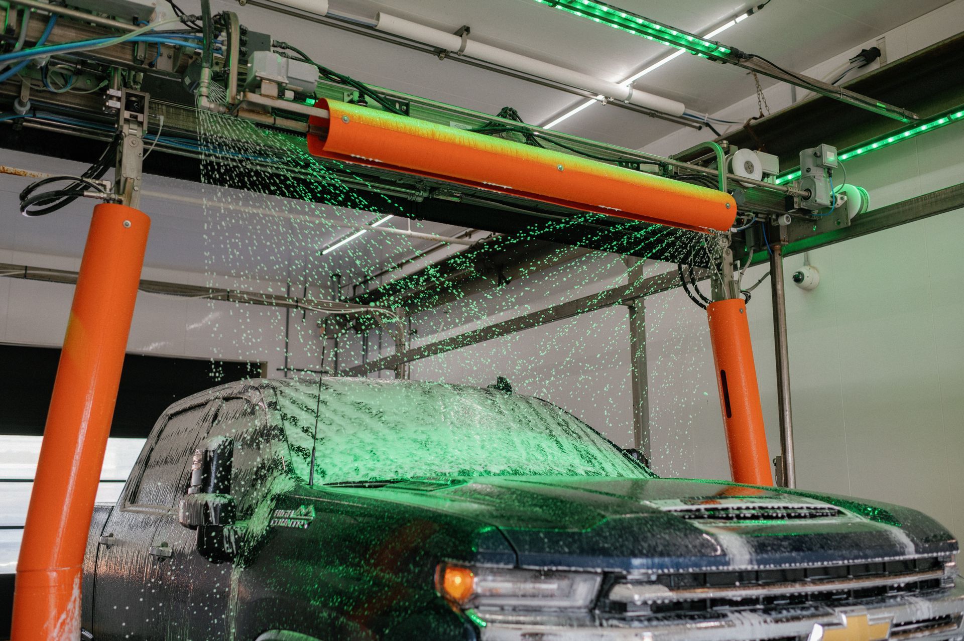 A dark-colored pickup truck being washed in a modern car wash with bright orange support pillars and green neon lighting.