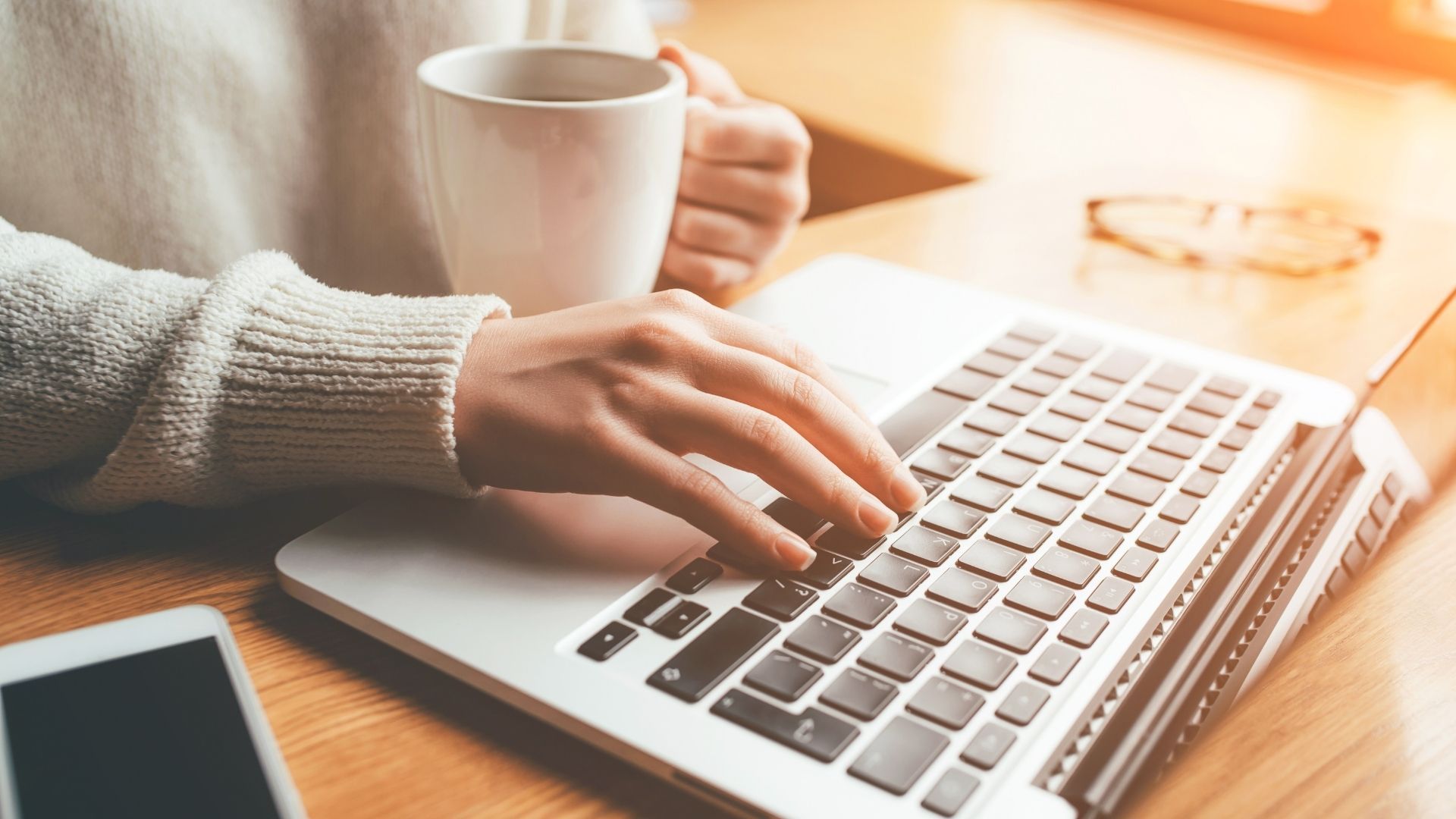 A person is typing on a laptop while holding a cup of coffee.