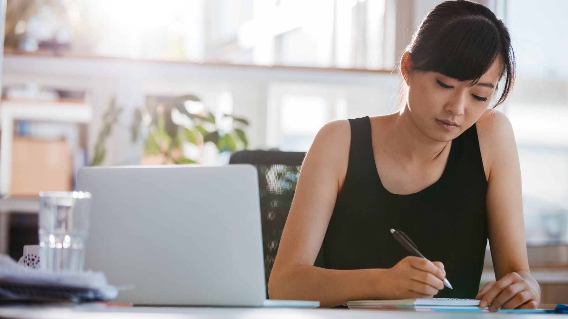 A woman is sitting at a desk with a laptop and writing on a piece of paper.