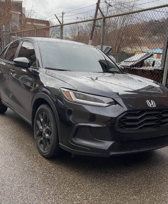 Black Honda HR-V parked on a wet street near a fence and buildings.