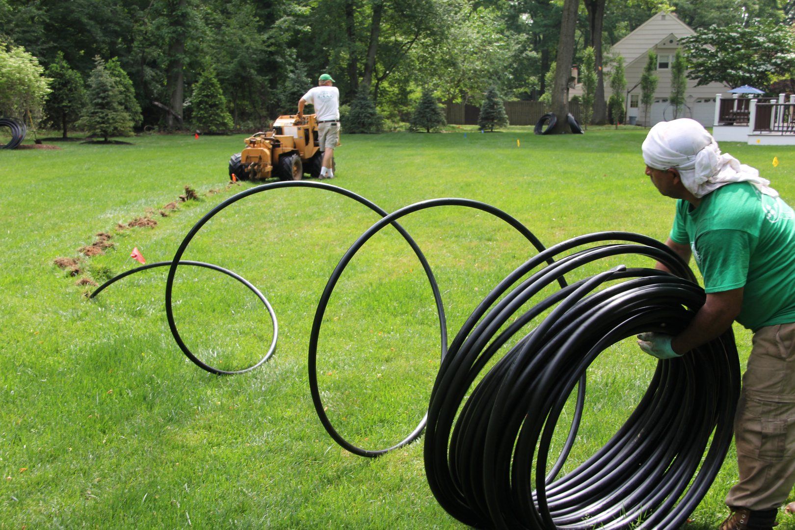 Man uncoils black tubing for yard irrigation, while another operates a stump grinder. Green lawn, sunny day.