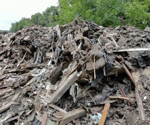 Pile of mixed debris including wood, asphalt, and other construction waste, set against a backdrop of green trees.