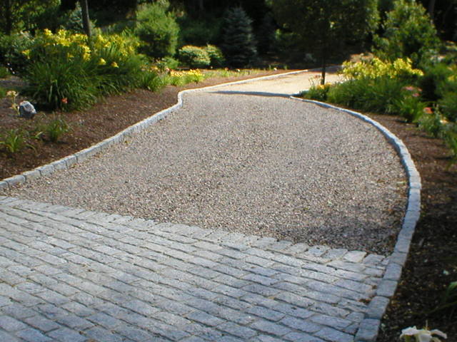 Gravel driveway bordered by stone, leading to a landscaped area with greenery.