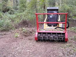 Red brush cutter attached to a yellow skid steer clears a path through brush and small trees.