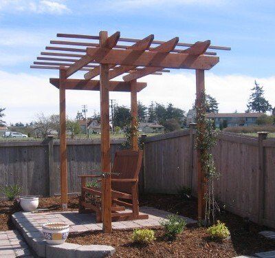 Wooden pergola with chair, in a backyard setting. Brown wood, blue sky, and a fence are visible.