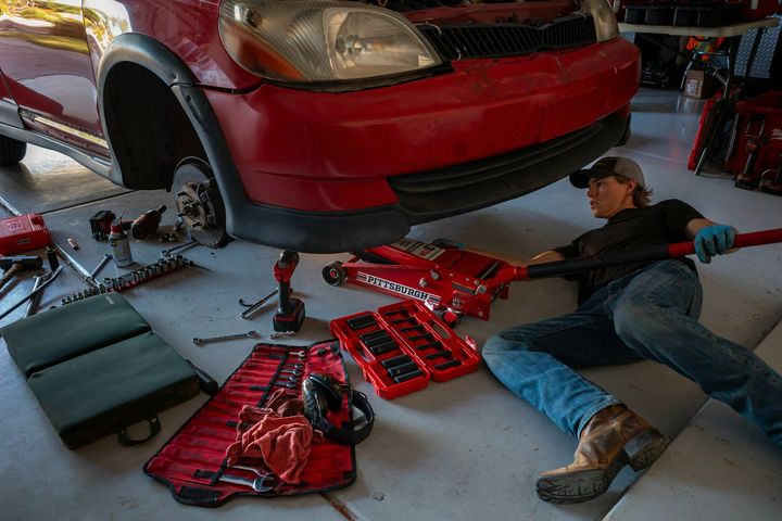 A mechanic wearing navy blue overalls works under the open hood of a car.