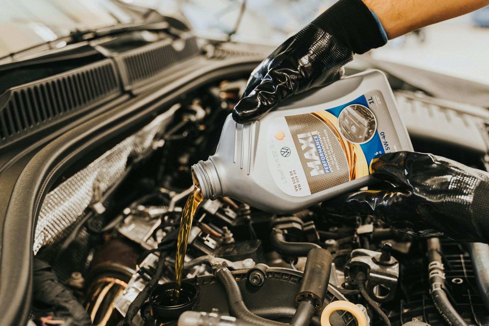A mechanic wearing black gloves pours golden motor oil into the engine compartment of a vehicle.