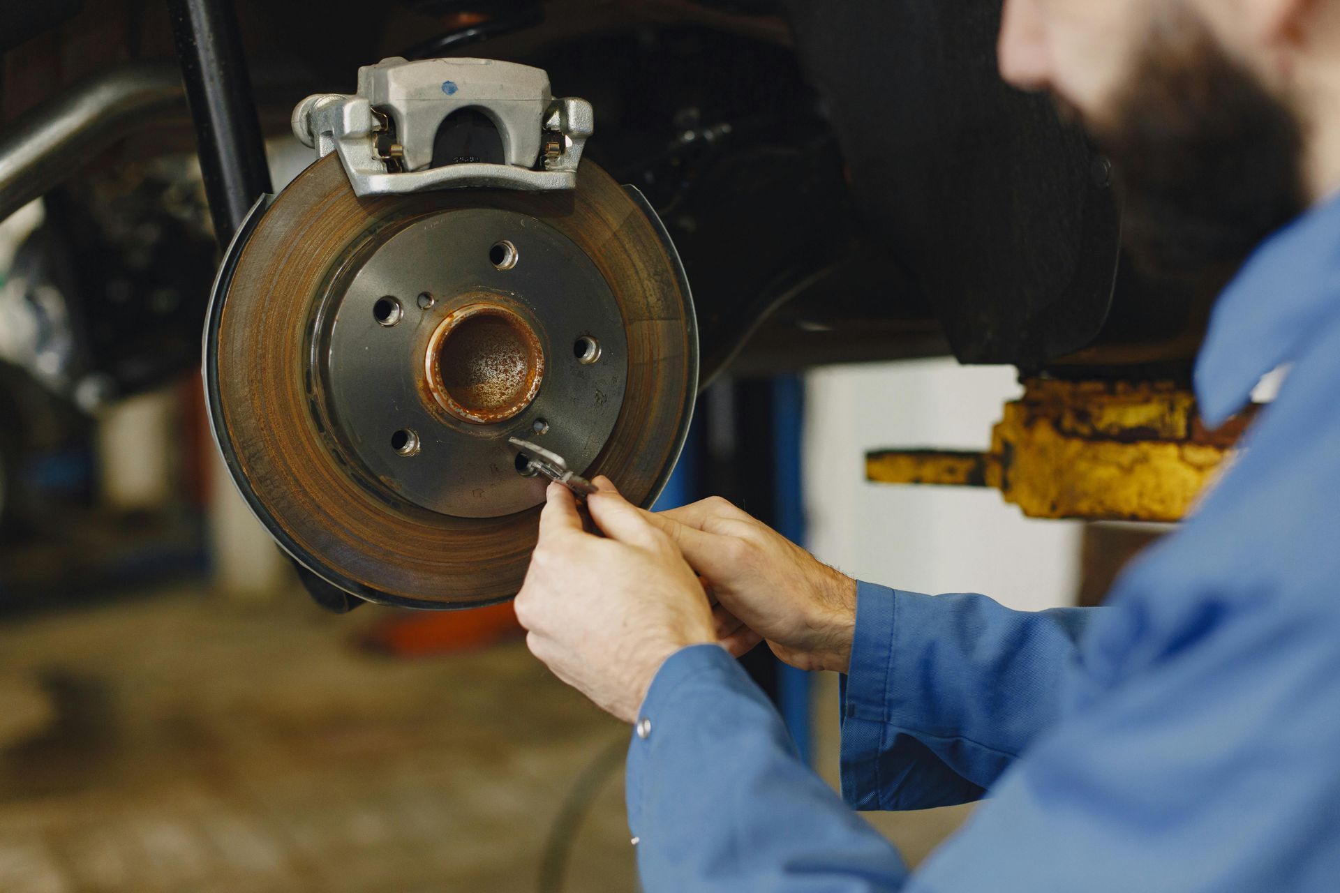 A mechanic in a blue shirt works on the rusted brake disc assembly of a vehicle in an auto repair shop.