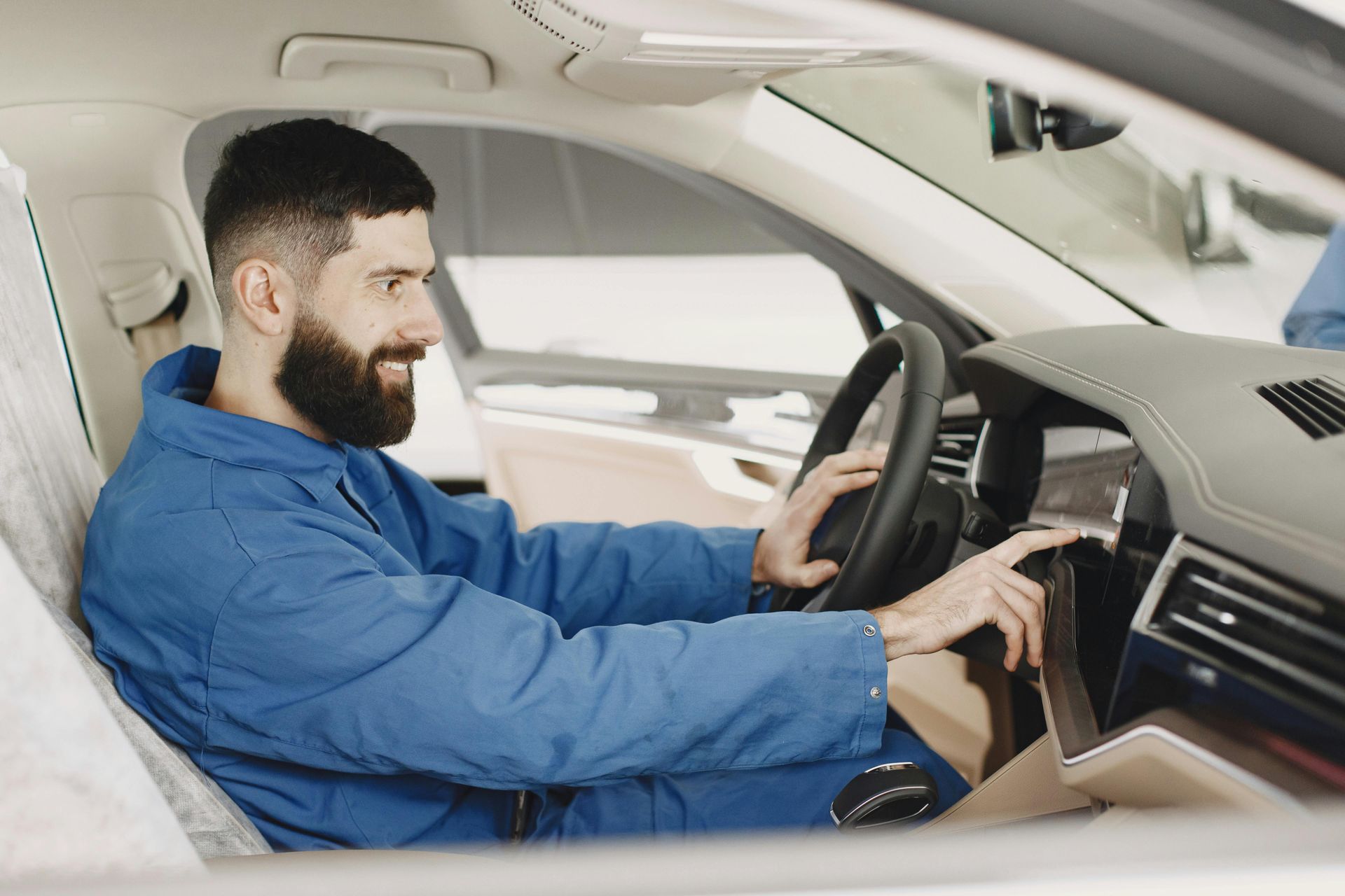 A bearded mechanic in a blue uniform sits in a car, testing the dashboard's touchscreen display.