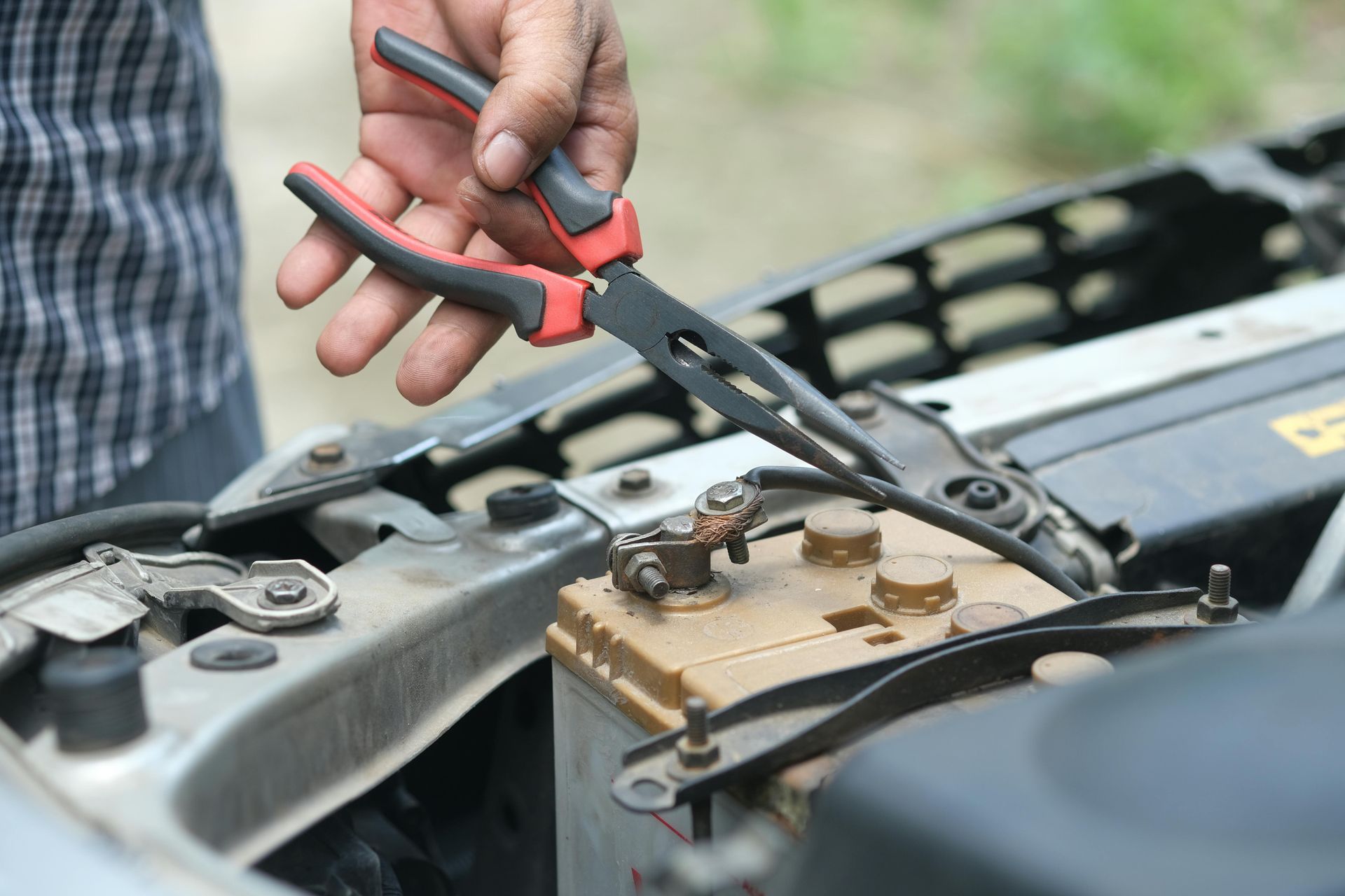 A person uses long-nose pliers to reach into an open car hood near the battery terminals.