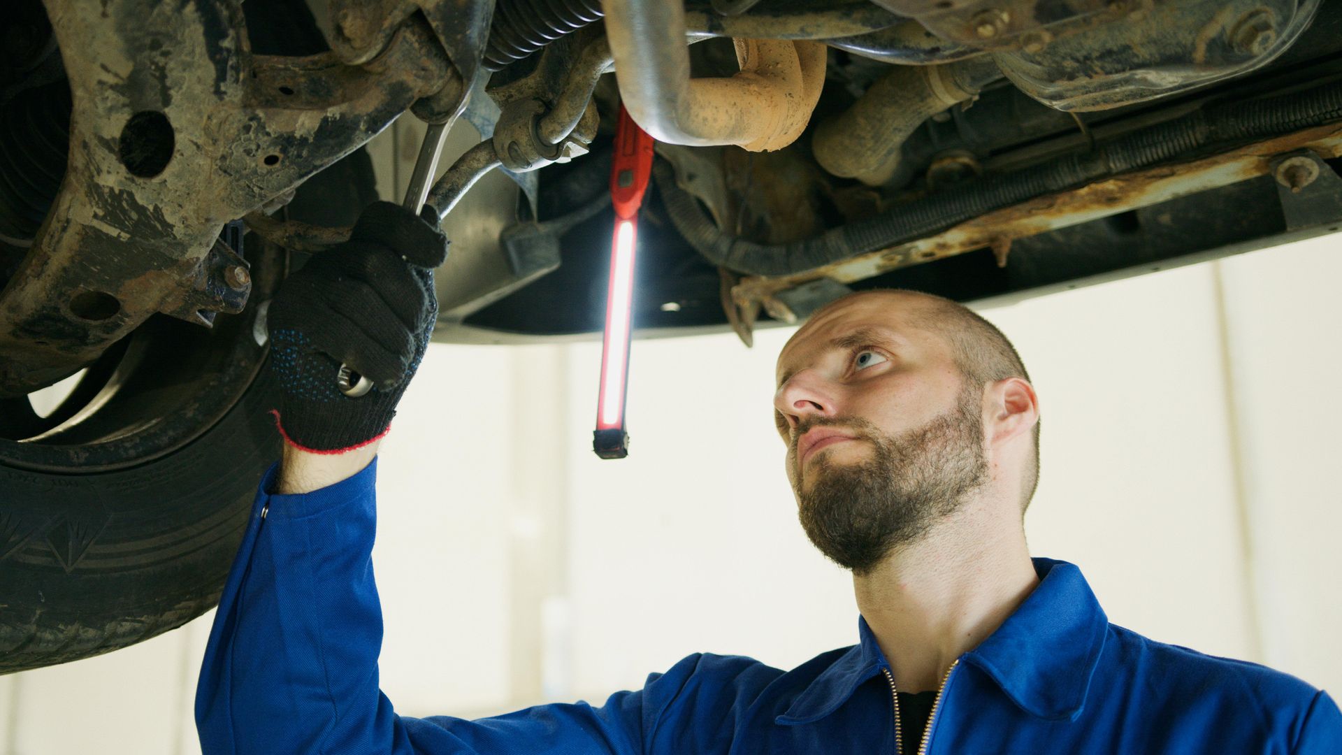 A mechanic in a blue uniform working under a lifted car, holding a tool near the suspension with a work light nearby.