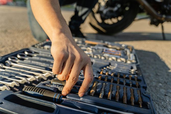 A hand selects a screwdriver from an open tool kit on the ground near a motorcycle.