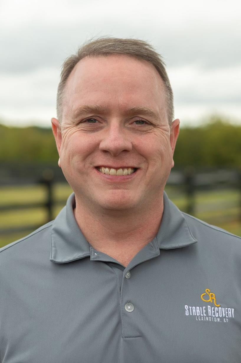 A woman with long brown hair is standing in front of a sign that says in k