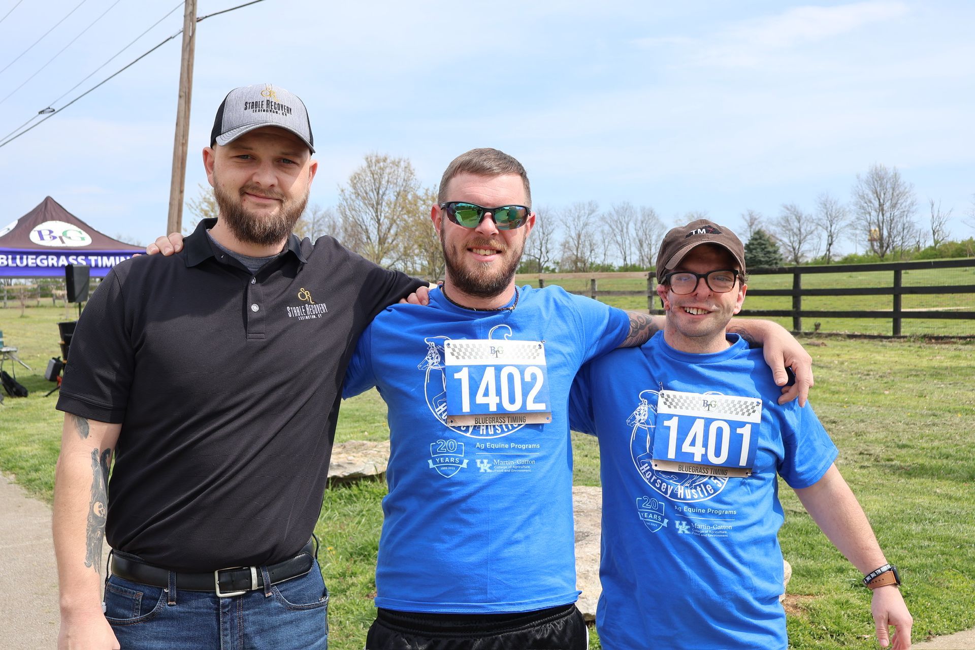 Three men in running shirts pose outdoors. Two have bibs, one with 