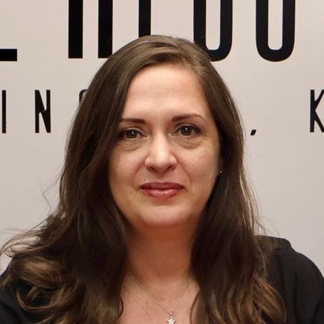 A woman with long brown hair is standing in front of a sign that says in k