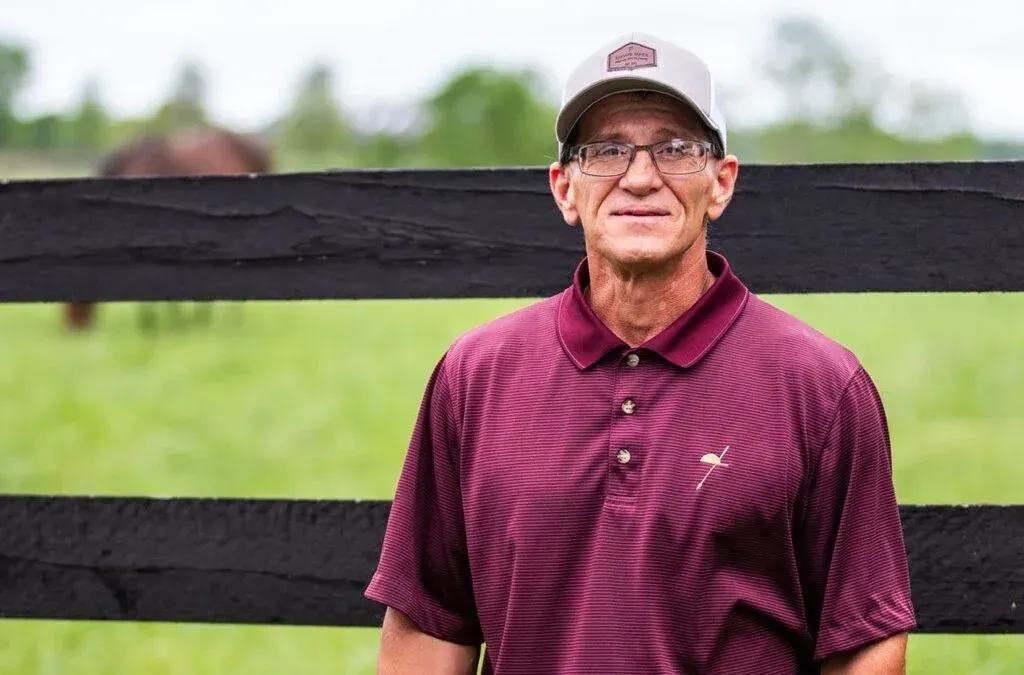 A man wearing a hat and glasses is standing in front of a fence in a field.