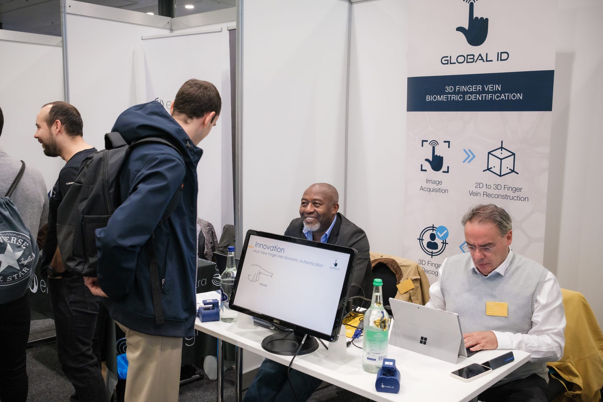 Trois personnes à un stand commercial, chacune avec un ordinateur portable et un écran ; l’une d’elles est assise et sourit derrière la table.