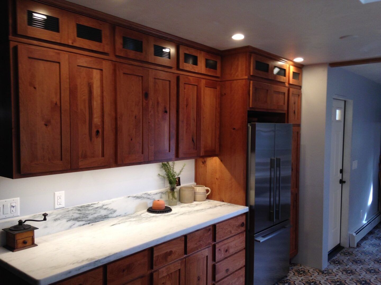 Kitchen with wooden cabinets, stainless steel refrigerator, white countertop, and under-cabinet lighting.
