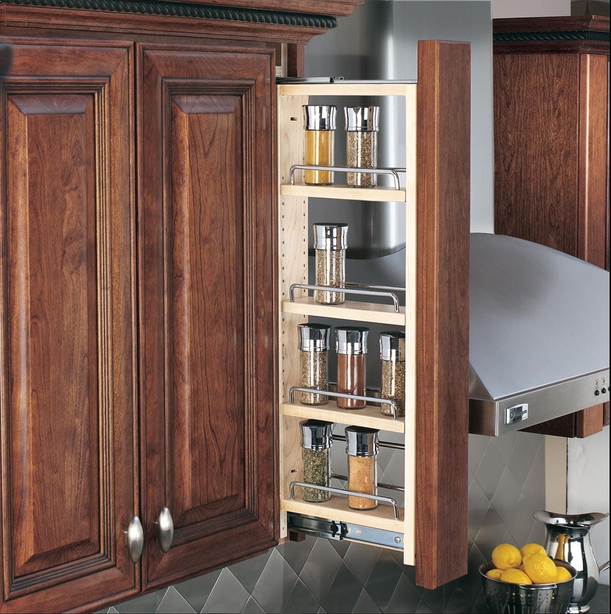 Pull-out spice rack next to dark wood kitchen cabinets, stainless steel range hood, and fruit bowl.