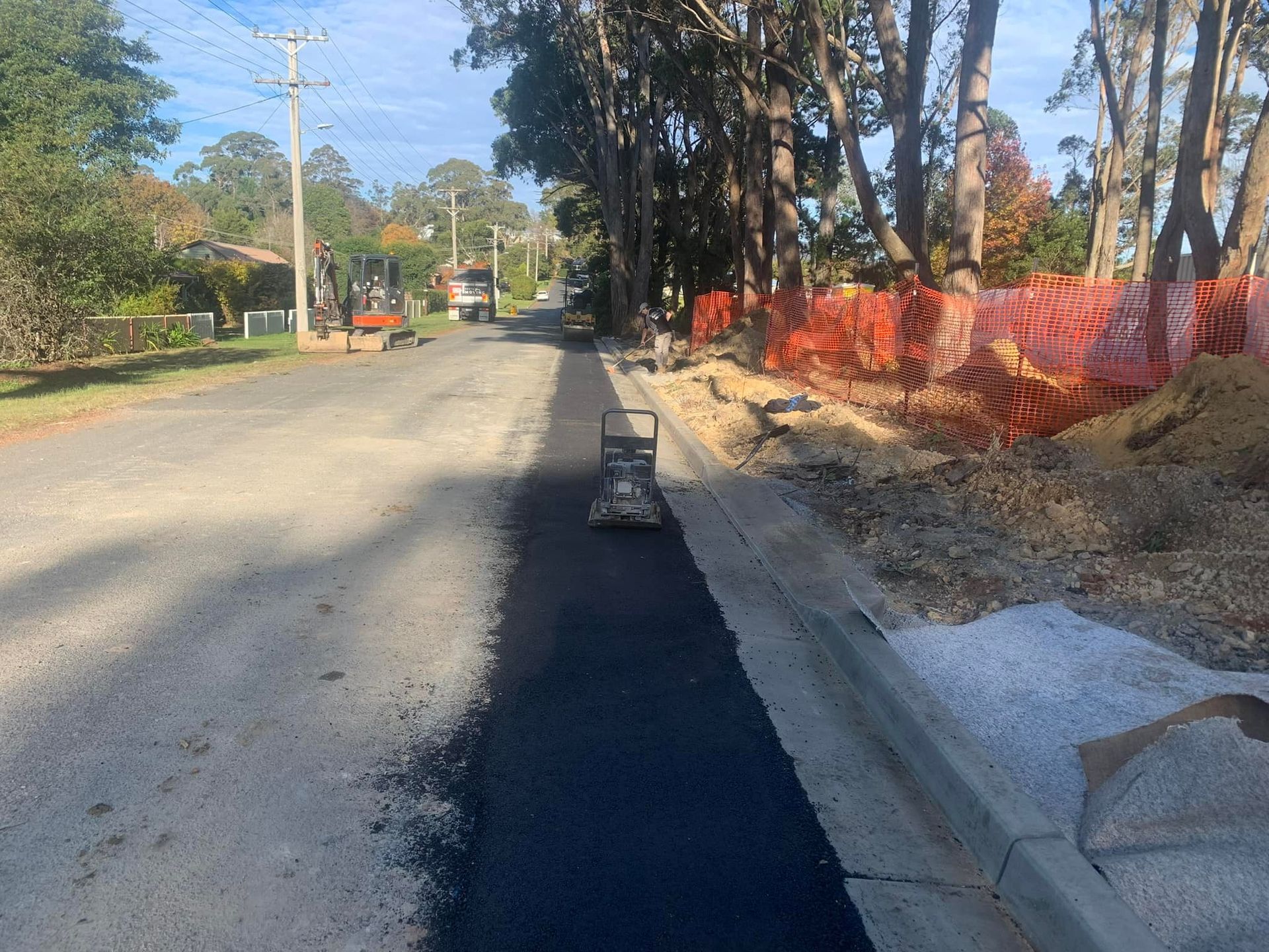 Residential street with new asphalt paving, curb, trees, and orange construction fencing on the right.