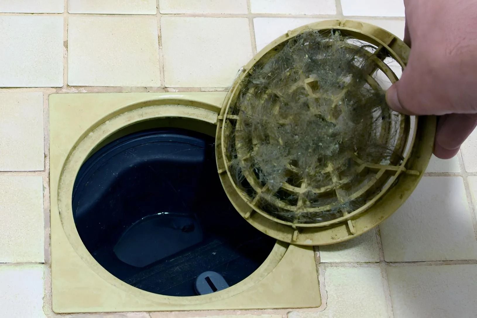 Hand lifting a dirty drain cover beside a dark blue circular drain in tiled floor