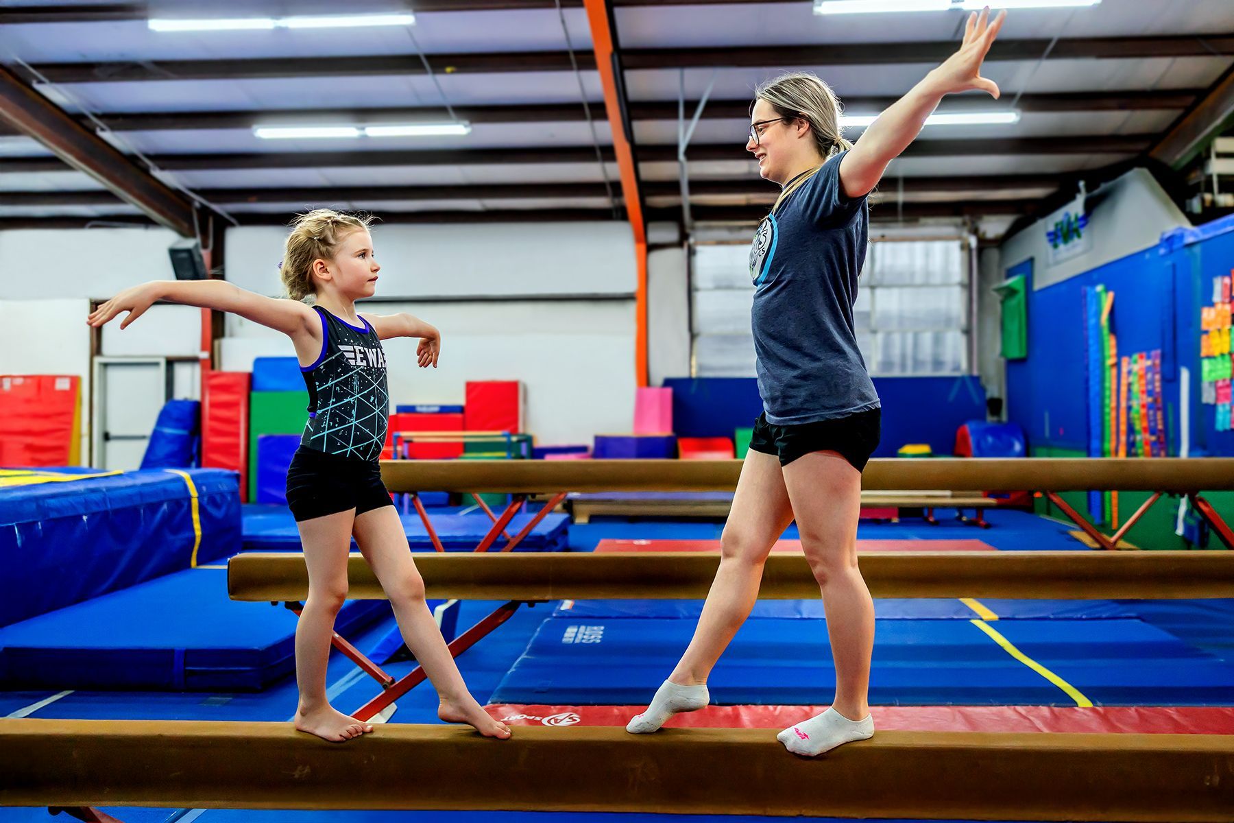 A woman and a little girl are standing on a balance beam in a gym.