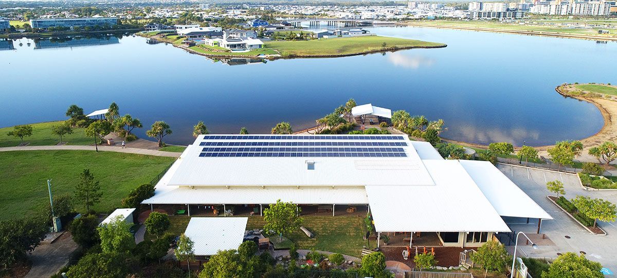 An aerial view of a building with solar panels on its roof, a lake, and a city in the background.
