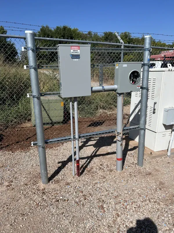 Electrical panel box mounted on a metal fence post, in a gravel setting.